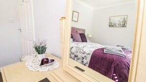 Une chambre avec un grand miroir, un lit recouvert d’une couvre-lit à motifs floraux, une table de chevet ornée d’un vase de fleurs et une porte.