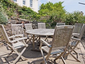 A round wooden table with six chairs, set on a stone patio with a stone wall and greenery in the background.