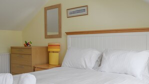 Un lit bien fait, avec du linge de lit blanc, une table de chevet en bois surmontée d’une lampe, une commode et un tableau encadré accroché au mur.