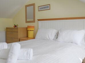 Un lit bien fait, avec du linge de lit blanc, une table de chevet en bois surmontée d’une lampe, une commode et un tableau encadré accroché au mur.