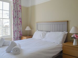 A neatly made bed with white linens, two bedside lamps, a wooden nightstand, and a window with floral curtains.