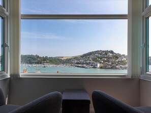 A view from a window overlooking a harbor with boats, a hill with buildings, and a chair in the foreground.