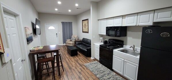 A compact kitchen with white cabinets, a black refrigerator, a stove, and a microwave. There is a wooden dining table with chairs and a sofa in the living area.