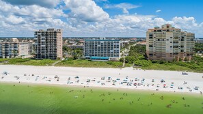 A beachfront hotel with multiple floors, a pool, and a beach area with umbrellas and people.