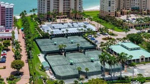 Un hotel su più piani con campo da tennis, circondato da palme e vista sulla spiaggia.