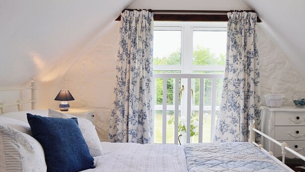 A bedroom with a bed, a nightstand, a lamp, a window with curtains, and a view of greenery outside.