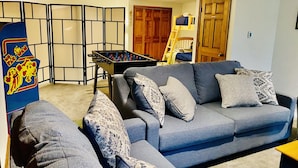 Un salon avec un canapé gris, des coussins à motifs, une table de baby-foot et une porte en bois.