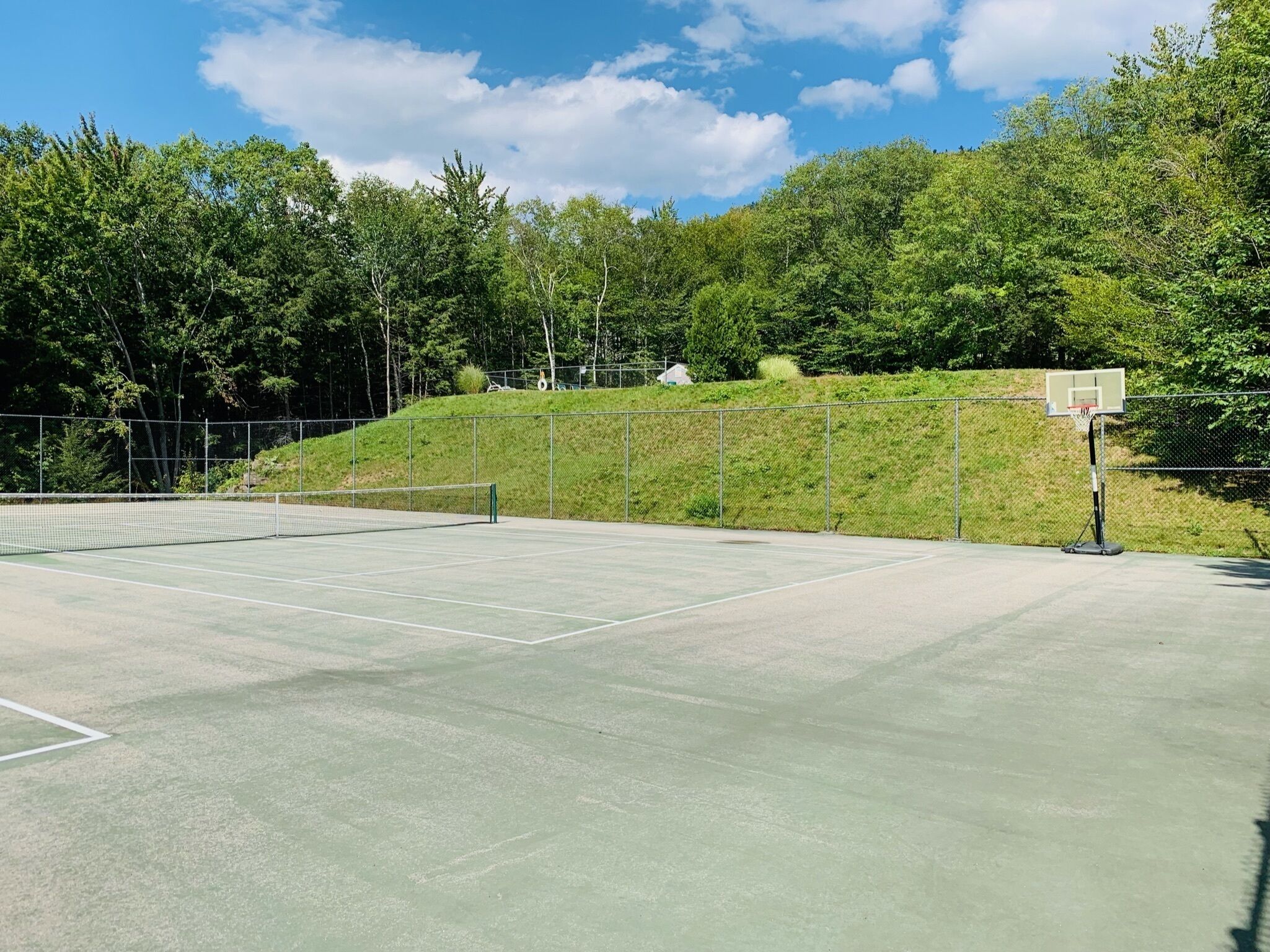 An outdoor tennis court with a net, surrounded by a chain-link fence and trees.