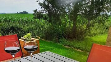 En picknickuppsättning med olika sorters ostar, frukter och nötter, arrangerat på en träbricka, placerad på ett utomhusbord med utsikt över en grön äng och träd i bakgrunden.