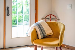 Un sillón color mostaza con una manta a rayas, situado frente a una ventana con vistas a la vegetación.