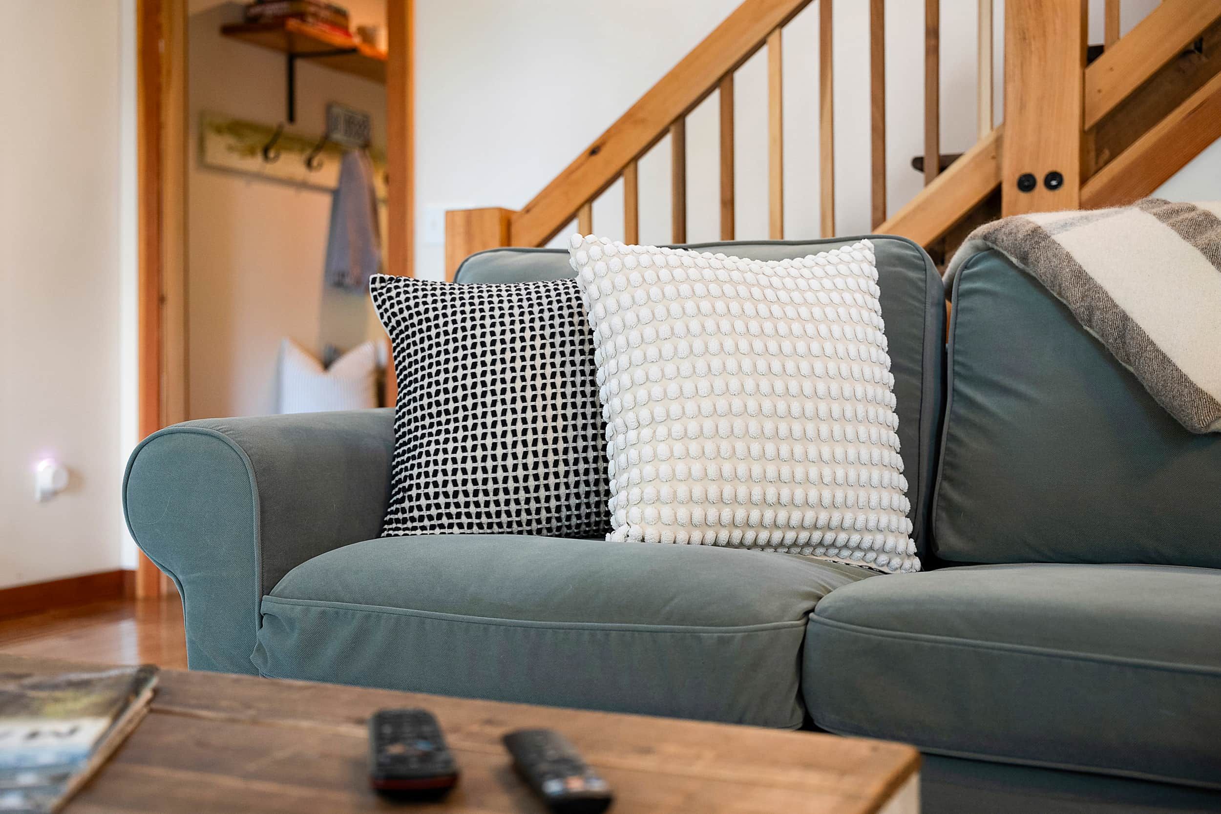 A teal sofa with patterned pillows, a wooden coffee table with remotes, and a staircase in the background.