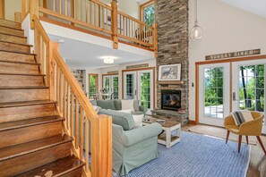 Un escalier en bois menant à une mezzanine, un salon confortable avec une cheminée et un mur de pierre orné d’une enseigne « Sunday River ».