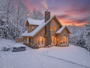 Een blokhut met twee verdiepingen en een stenen schoorsteen, omgeven door met sneeuw bedekte bomen.