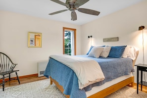 Un dormitorio con cama, ventilador de techo, ventana, silla y mesita de noche.