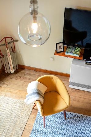 Una habitación con un sillón amarillo, una lámpara colgante grande, un televisor de pantalla plana y una estantería con libros y un cuadro enmarcado.
