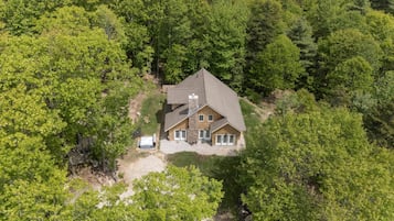 An aerial view of a house surrounded by dense forest, with a visible driveway and a small patio area.