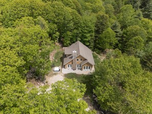 An aerial view of a house surrounded by dense forest, with a visible driveway and a small patio area.