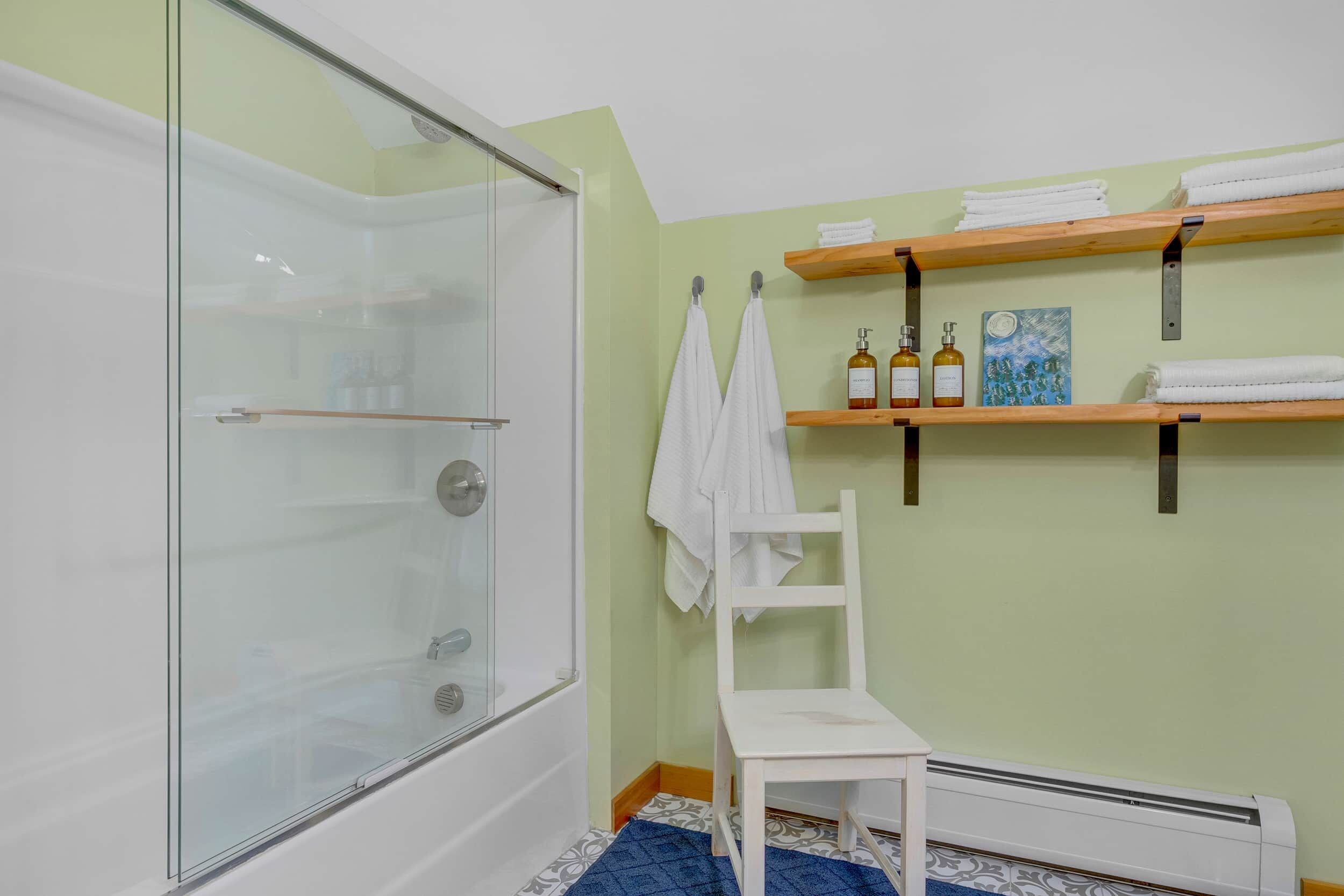 A bathroom with a glass shower enclosure, a white chair, and shelves with towels and toiletries.