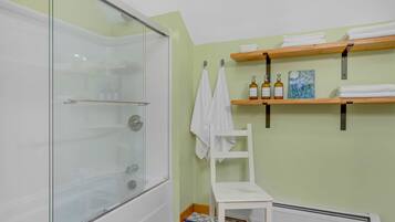 A bathroom with a glass shower enclosure, a white chair, and shelves with towels and toiletries.