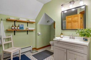 Et badeværelse med en træstol, en vask med et spejl, hylder med toiletartikler og et mønstret gulv.
