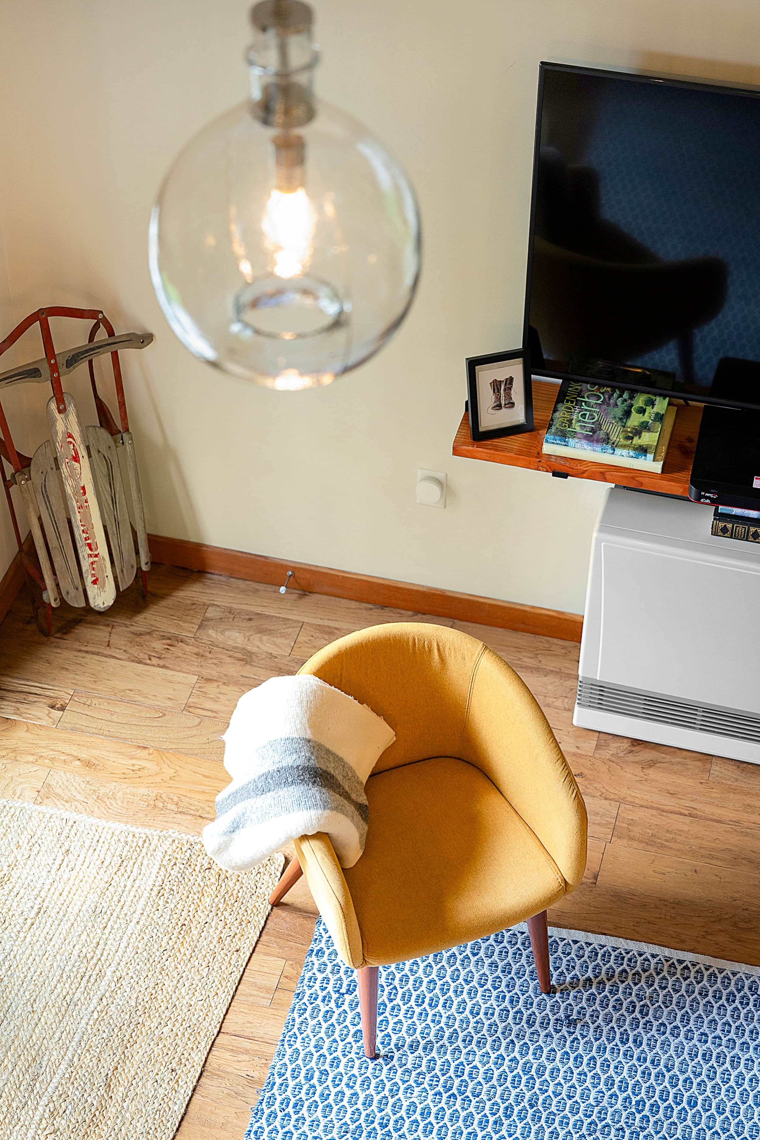 Una habitación con un sillón amarillo, una lámpara colgante grande, un televisor de pantalla plana y una estantería con libros y un cuadro enmarcado.