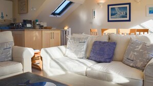 A cozy living room with a sofa, chairs, and a coffee table. There is a skylight above the sofa and a kitchen area in the background. - 3 bedroom accommodation in Buckley St (Buckley St)