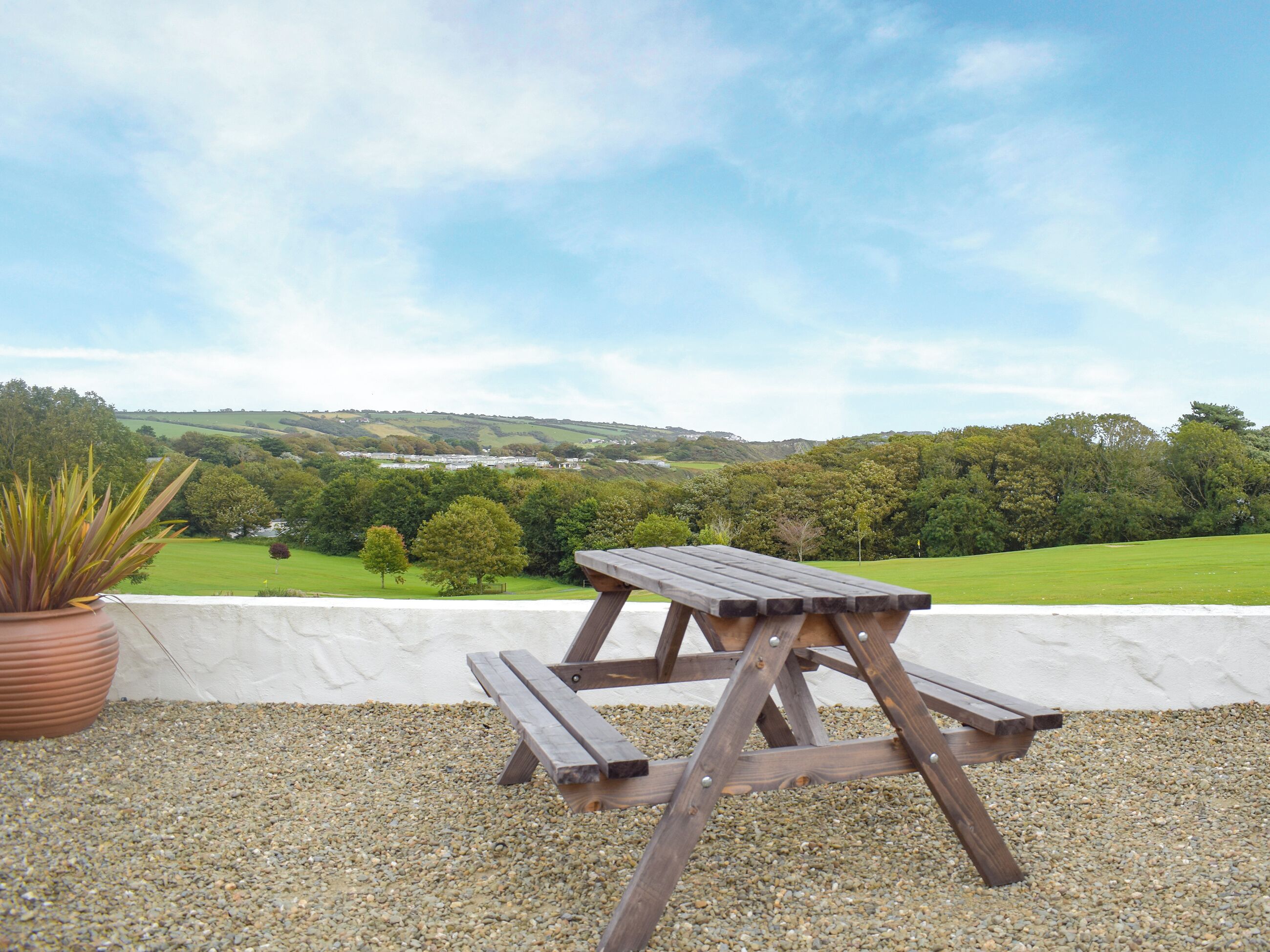 Una mesa de picnic de madera con bancos sobre un terreno de grava, una planta en maceta a la izquierda y una vista panorámica de campos verdes y colinas al fondo.