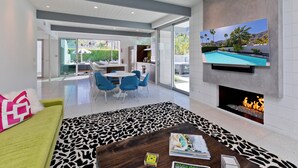 Ein modernes Wohnzimmer mit Kamin, Fernseher mit Blick auf den Pool und einem Essbereich mit blauen Stühlen.