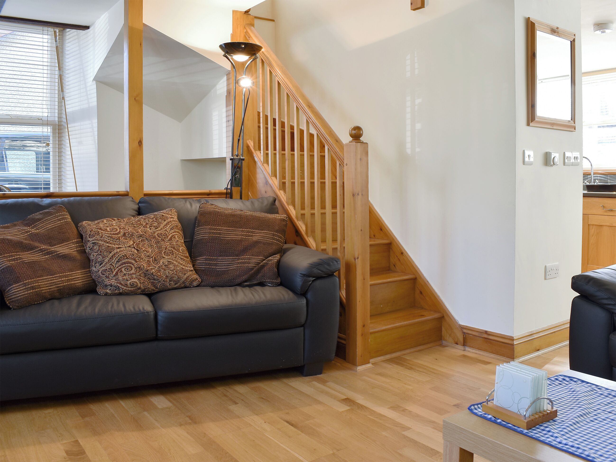 A modern living room with a dark leather sofa, patterned cushions, a wooden staircase, and a kitchen area in the background.