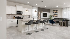 A modern kitchen with white cabinets, a central island with bar stools, stainless steel appliances, pendant lighting, and a grey sofa in the living area. - Dream Residence Equipped with a Game Room, Snooker Table, PlayStation 5 (Davenport)