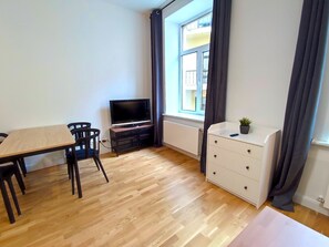 A small, tidy room with a wooden floor, a TV on a stand, a dining table with chairs, a white dresser, and a window with curtains.