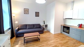 En moderne stue med sofa, sofabord, et lille sidebord med en plante, og et køkkenområde med skabe og komfur.
