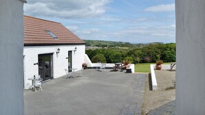 A white building with a red-tiled roof, a patio with tables and chairs, and a view of green hills and a distant coastline. - Gardener's Cottage - UK6713 (Lydstep, near Tenby)