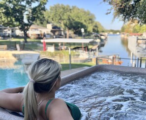 A person relaxing by a hot tub with a view of a river and buildings in the background.