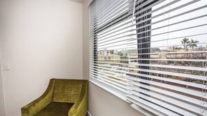 Una habitación con un sillón verde, una ventana con persianas horizontales y vistas a edificios y árboles.