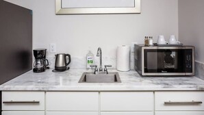 Una cocina con lavamanos, cafetera, hervidor, microondas y un cuadro en la pared.