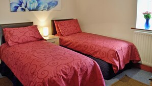 A bedroom with two beds, a nightstand, a lamp, a window with curtains, and a floral painting on the wall. - 1 bedroom accommodation in Belford near Bamburgh (Belford near Bamburgh)