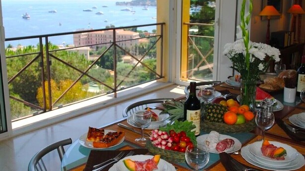 Una mesa de comedor dispuesta con diferentes platos, una botella de vino y vistas al mar y edificios.