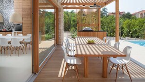 An outdoor dining area with a wooden table and white chairs, a grill, and a pool in the background. - Awarded Design Villa Olea, Luxury Villa with heated pool & Seaview (Klimno (Krk))