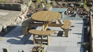 A wooden outdoor seating area with a round table and benches, surrounded by a stone patio and a garden with rocks and plants. - 2 bedroom accommodation in Troutbeck Bridge (Troutbeck Bridge)