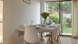 Un coin repas avec une table dressée pour quatre personnes, des chaises blanches, un vase rempli de fleurs et une vue sur la verdure par une grande fenêtre.