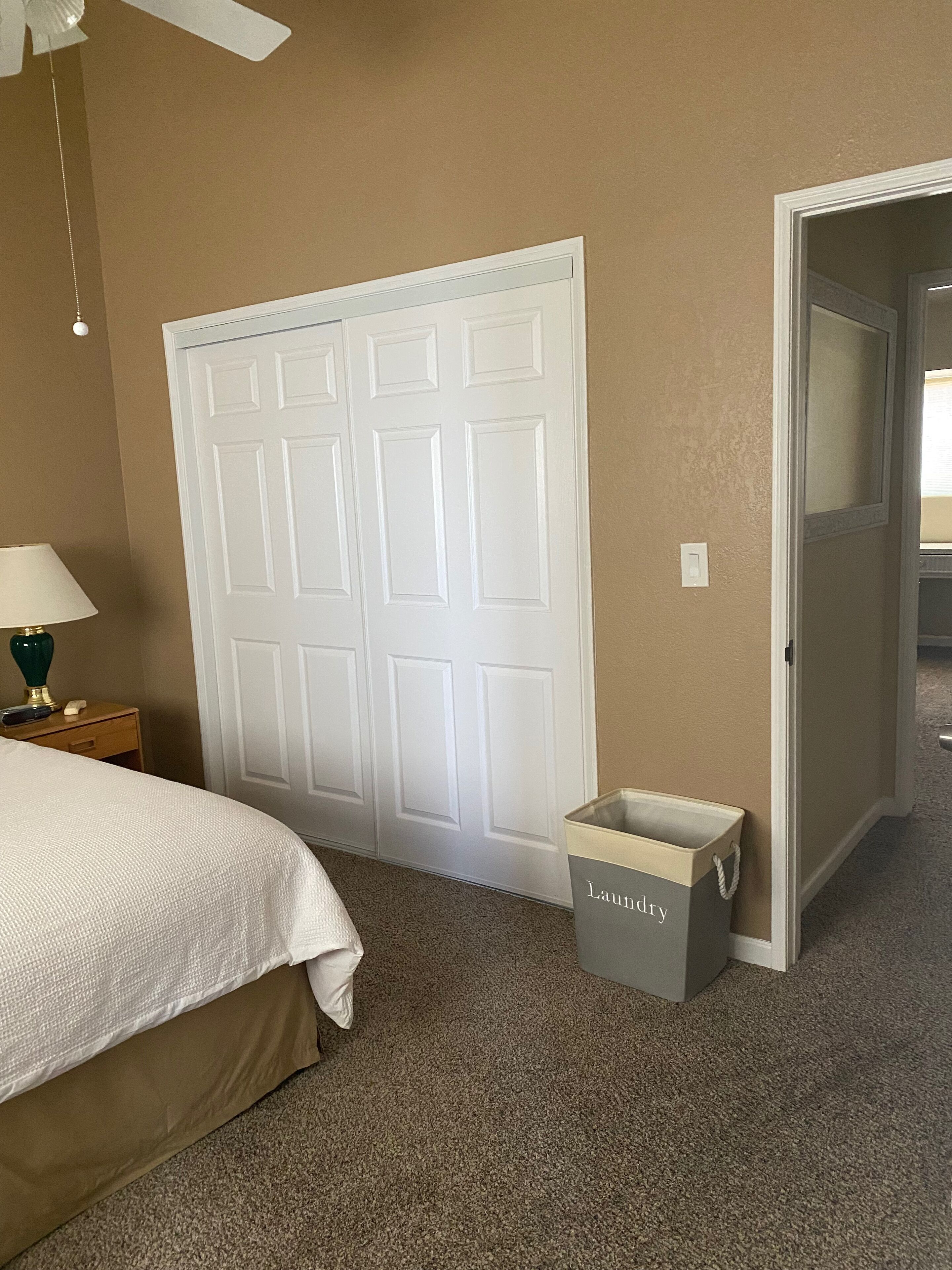 A bedroom with a bed, a nightstand, a lamp, a ceiling fan, a door, and a laundry basket.