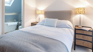 A bedroom with a bed, two bedside tables, a nightstand, a lamp, a window, and a bathroom visible through an open door.