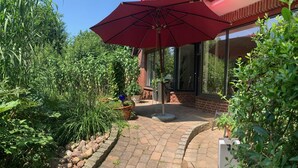 Un patio con sombrilla roja, plantas en macetas y un sendero de ladrillo que lleva a una puerta de cristal.