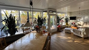 Amplia sala de estar con una gran mesa de comedor de madera, múltiples sillas, un sofá y un televisor montado en la pared. Varias plantas en macetas y un ventanal con vistas al exterior.