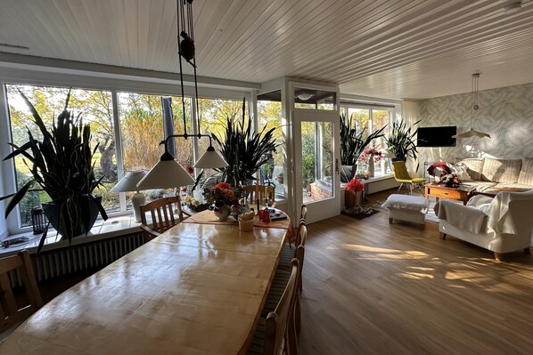 Un vaste salon comprenant une grande table à manger en bois, plusieurs chaises, un canapé et un téléviseur fixé au mur. On y trouve également plusieurs plantes en pot et une fenêtre donnant sur l’extérieur.