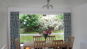 Un comedor con una mesa, sillas y un cuenco de fruta. Se ve una ventana con cortinas y vistas al exterior con vegetación.
