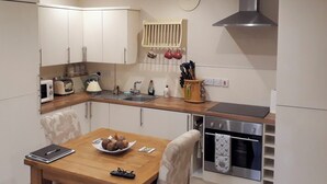 Cocina compacta con armarios blancos, una mesa de comedor de madera, horno empotrado y un reloj en la pared.