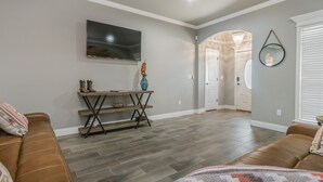 Een ruime woonkamer met een grote flatscreen televisie aan de muur, een houten console met decoratieve objecten, en een gezellige bruine leren bank.