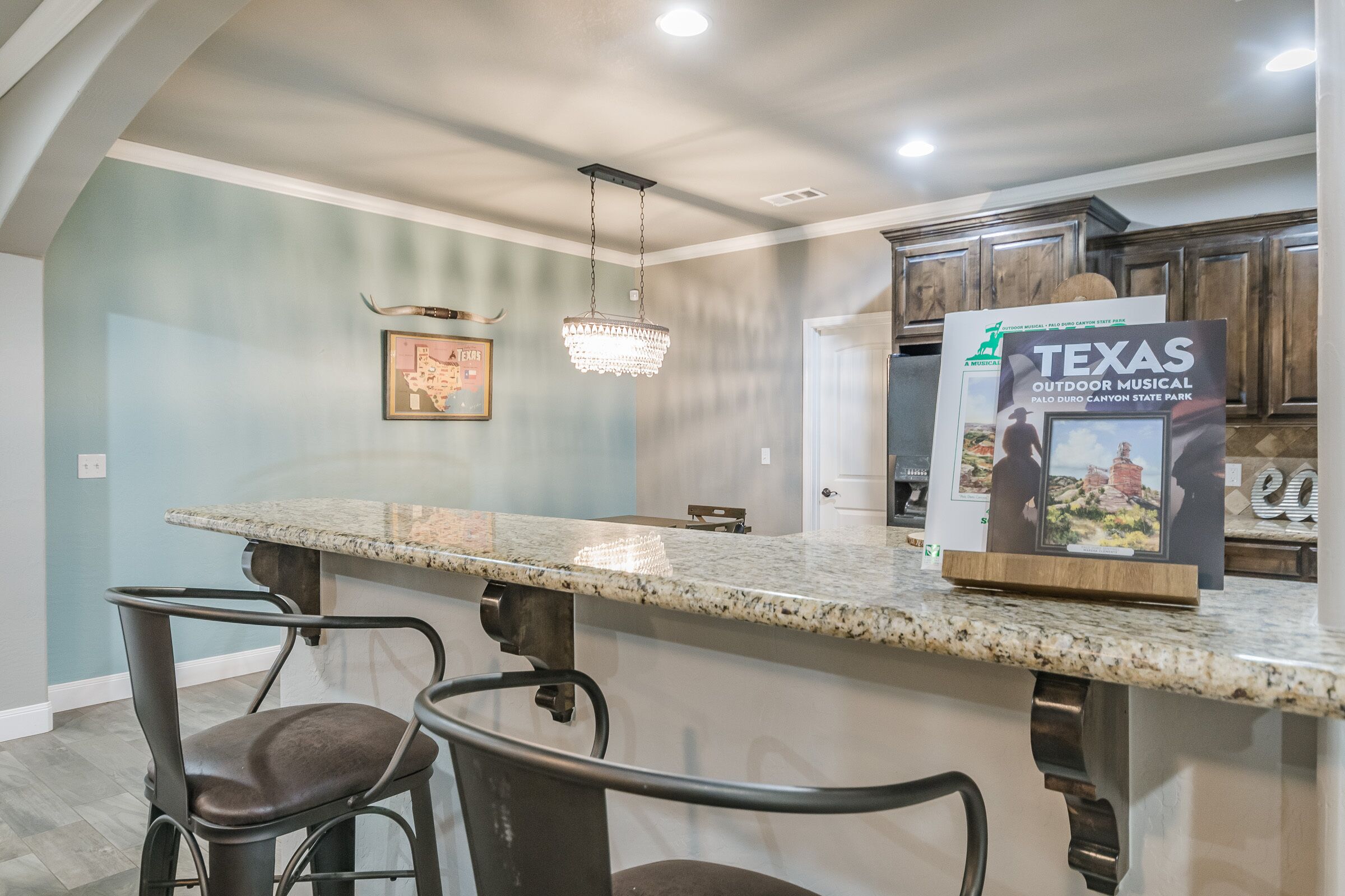 A kitchen with granite countertops, bar stools, a chandelier, and a Texas Outdoor Musical sign.